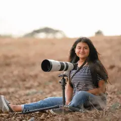 Młoda kobieta z długimi włosami siedzi na ziemi w polu, obok niej potężny teleobiektyw na statywie. Kompozycja otwarta podkreśla jej pasję do fotografii przyrodniczej.