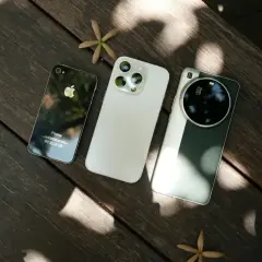 Trzy telefony do zdjęć: stary iPhone, nowy iPhone i smartfon z aparatem Leica, leżące na drewnianym stole.
