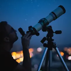 Mężczyzna obserwuje nocne niebo przez teleskop na tle rozświetlonego miasta.