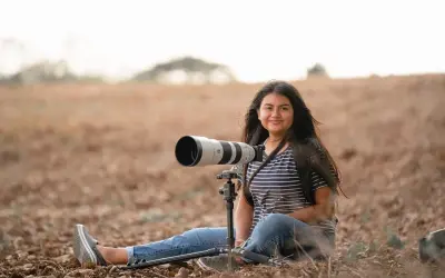 Młoda kobieta z długimi włosami siedzi na ziemi w polu, obok niej potężny teleobiektyw na statywie. Kompozycja otwarta podkreśla jej pasję do fotografii przyrodniczej.