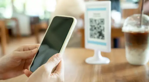 Ręce trzymają telefon z Androidem, gotowe do zeskanowania kodu QR na stoliku w kawiarni.