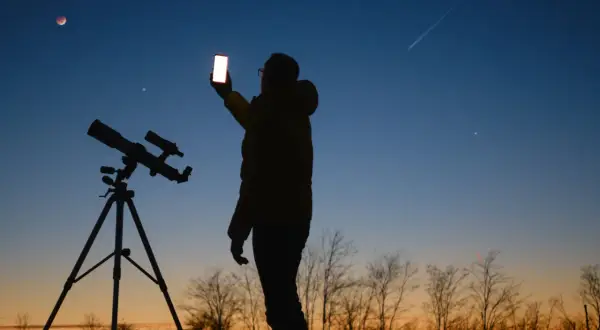 Sylwetka osoby z telefonem i teleskopem na tle nieba z zaćmieniem Księżyca i smugą po samolocie.