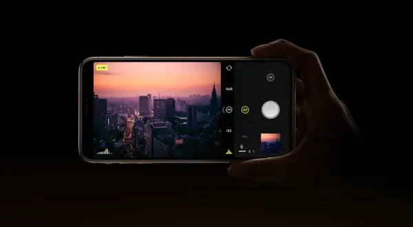 Dłoń trzyma smartfon z widokiem na miasto o zachodzie słońca. Na ekranie widać interfejs aparatu, gdzie można ustawić ISO.