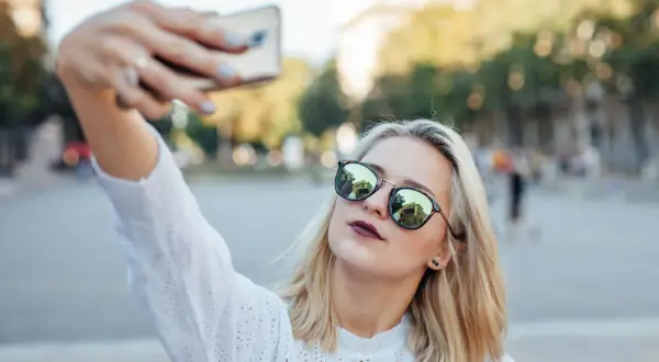 Blondynka w okularach robi sobie selfie telefonem. W okularach odbija się budynek. Oto jak zrobić dobre selfie telefonem.
