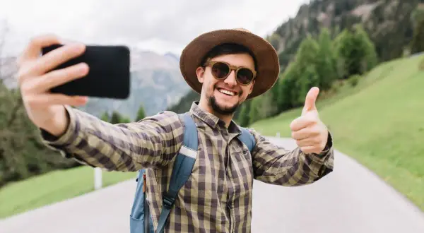 Uśmiechnięty turysta w kapeluszu robi sobie selfie z kciukiem w górę na tle gór. Widać, jak z łatwością zrobić selfie.